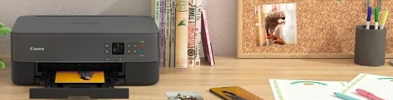 Canon-skrivare på skrivbord som skriver ut ett färgfoto, omgiven av foton, mobil och kontorsmaterial